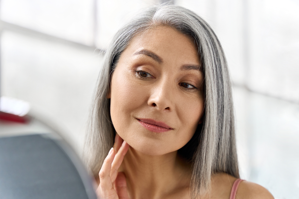 Portrait of gorgeous happy middle aged mature asian woman, senior older 50s lady pampering touching face looking at herself at mirror indoors.