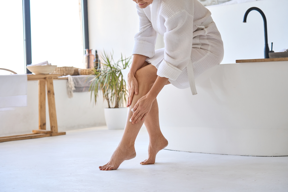 Mid age adult 50s age mature woman applying varicose prevention treatment cream massaging legs sitting on bathtub wearing white bathrobe